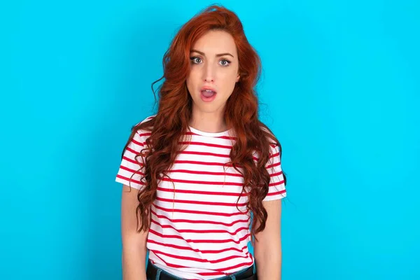 Oh my God. Surprised Young caucasian redhead woman wearing striped t-shirt over blue background stares at camera with shocked expression exclaims with unexpectedness,