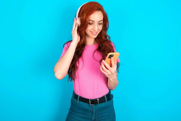 Happy Young caucasian redhead woman wearing pink t-shirt over blue background feels good while focused in screen of smartphone. People, technology, lifestyle