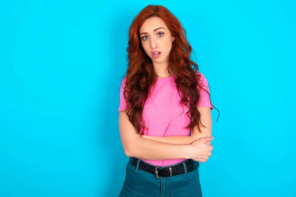 Shocked embarrassed Young caucasian redhead woman wearing pink t-shirt over blue background keeps mouth widely opened. Hears unbelievable novelty stares in stupor