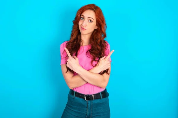 Young caucasian redhead woman wearing pink t-shirt over blue background  crosses arms and points at different sides hesitates between two items or variants. Needs help with decision