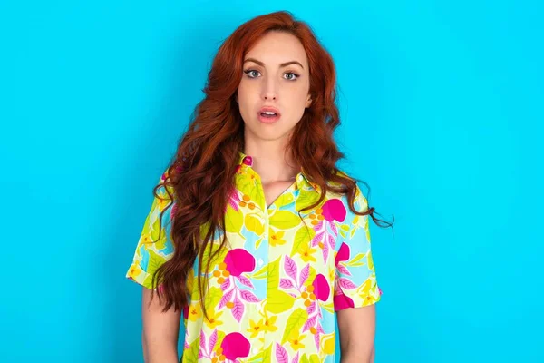 Shocked Young caucasian redhead woman wearing colorful shirt over blue background stares bugged eyes keeps mouth opened has surprised expression. Omg concept