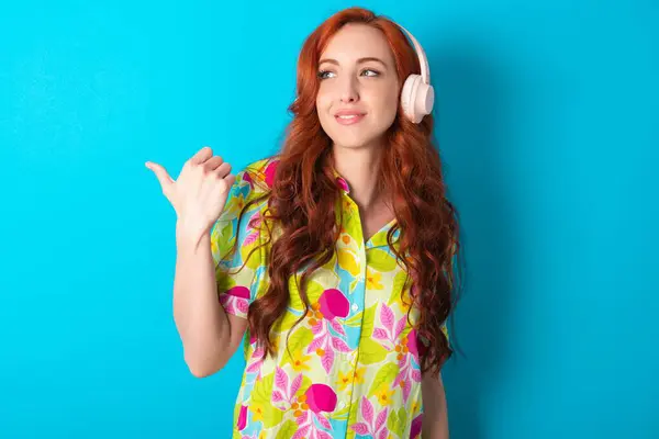 stock image Young caucasian redhead woman wearing colorful shirt over blue background listens audio track via wireless headphones points thumb away advertises copy space