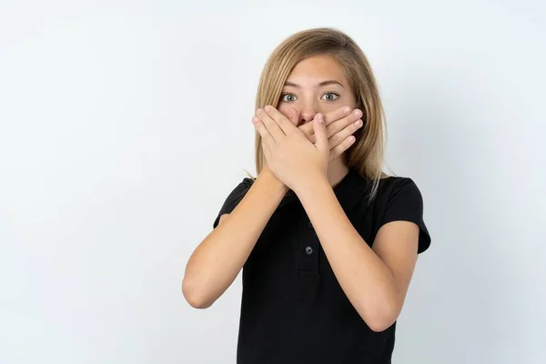 白いスタジオの背景に黒いTシャツを着た美しい10代の女の子がミスのために手で口をカバーショックを受けました 秘密概念 — ストック写真