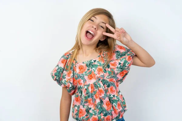 美しい10代の女の子が白いスタジオの背景に花のTシャツを着て顔の上に指で平和のシンボルを行います 明るい勝利を示す笑顔 — ストック写真
