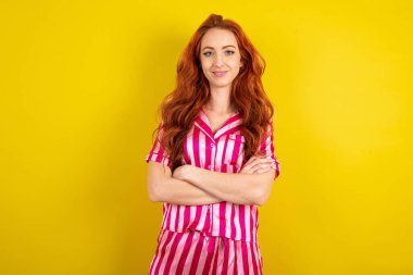 Rüya gibi bir istirahat, kızıl saçlı, pembe pijama giyen, sarı stüdyo arka planında kollarını kavuşturan genç bir kadın.,
