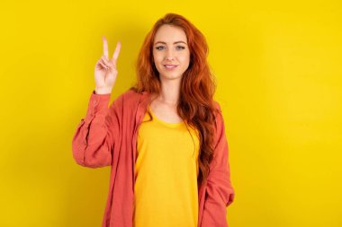 Sarı stüdyo arka planında duran genç, güzel kızıl saçlı kadın iki numaralı parmaklarıyla gülümseyerek kendinden emin ve mutlu bir şekilde onu işaret ediyor..