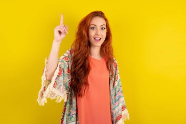 Genç, güzel, kızıl saçlı bir kadın sarı stüdyo arka planında duruyor. Parmakla işaret ediyor ve dahiyane düşüncelerden esinlenerek bakıyor. İyi bir fikir işareti gösteriyor.