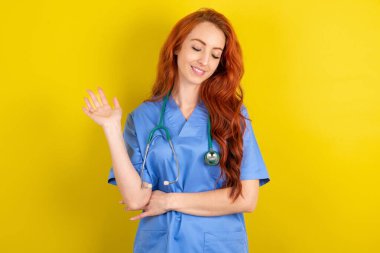 Aşırı neşeli, genç, kızıl saçlı doktor kadın sarı stüdyo arka planında avucunu kaldırıyor ve arkadaşları tarafından eğlendirilirken gözlerini kapıyor.