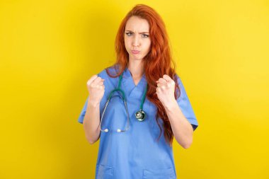 Displeased annoyed young red-haired doctor woman over yellow studio background clenches fists, gestures pissed, ready to revenge, looks with aggression at camera stands full of hate, being pressured