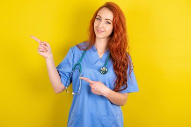 İyimser, genç, kızıl saçlı doktor kadın sarı stüdyo arka planında iki eliyle boş bir yere bakıyor..
