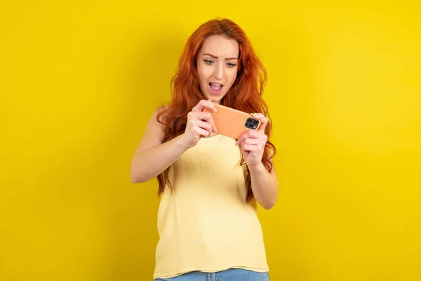 興奮した若い赤髪の肖像画は 携帯電話でゲームをプレイ黄色のTシャツ黄色のスタジオの背景を着て — ストック写真