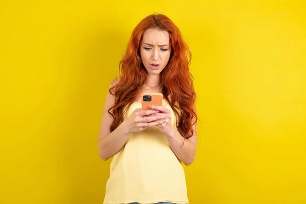 黄色のTシャツを着た若い赤髪の若者は スマートフォンのソーシャルメディアのニュースや重要な電子メールを読んで黄色のスタジオの背景を驚かせた — ストック写真