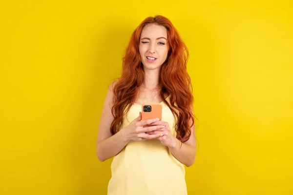 黄色のTシャツを着て興奮した若い赤髪黄色のスタジオの背景ウィンクと目は スマートフォンの使用を保持ソーシャルネットワークニュースを読む — ストック写真