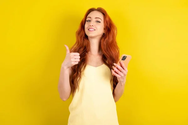 黄色のTシャツを着て若い赤髪の肖像黄色のスタジオの背景を使用して 大きな笑顔で幸せなスマートフォンでテキストメッセージ を署名を行う 指で親指を立てる 優れたサイン — ストック写真
