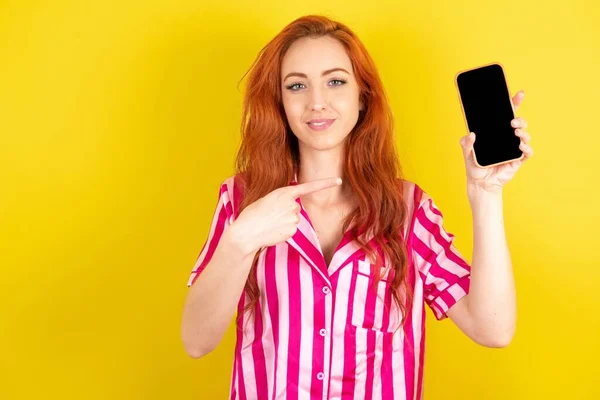 笑顔黄色のスタジオの背景にピンクのパジャマを着た若い赤い髪の女性モックアップコピースペース 空白の画面を持つ携帯電話上のインデックス指を指す — ストック写真