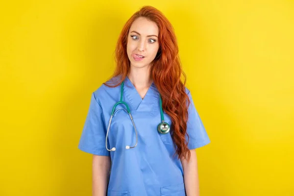 Funny young red-haired doctor woman over yellow studio background makes grimace and crosses eyes plays fool has fun alone sticks out tongue.