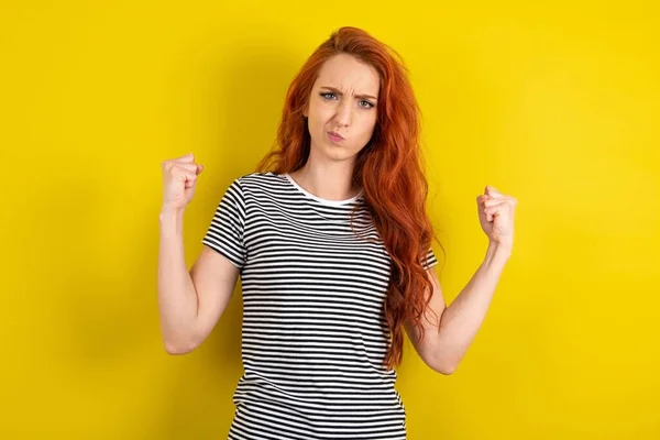Irritated young red-haired wearing striped t-shirt yellow studio background blows cheeks with anger and raises clenched fists expresses rage and aggressive emotions. Furious model