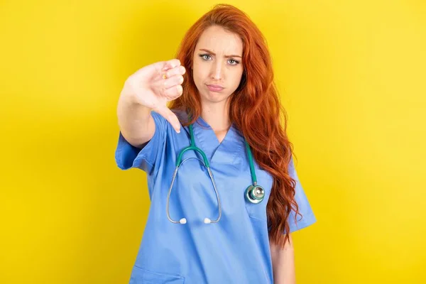 黄色のスタジオを背にした若い赤髪の医師の女性は 親指ダウンジェスチャーで拒絶と否定を示す不満と怒りを見ています 悪い表現 — ストック写真