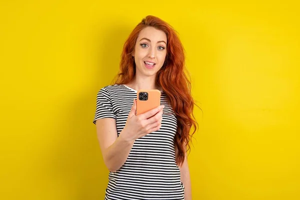若い赤い髪を身に着けているストライプのTシャツ黄色のスタジオの背景は手で携帯電話を保持し 肯定的なニュースを喜び 現代の携帯電話を使用して — ストック写真