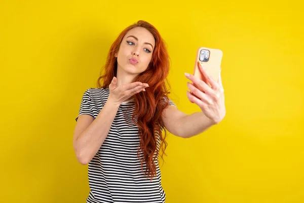若い赤い髪を身に着けているストライプのTシャツ黄色のスタジオの背景は スマートフォンのカメラでエアキスを吹いて 自撮りを取り オンラインコールを介してMwahを送信します — ストック写真