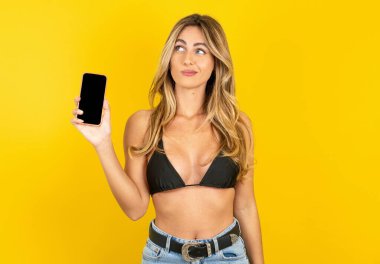 Sarı stüdyo arka planında bikini giyen genç sarışın kadın yeni cep telefonu tutuyor ve gizemli görünüyor. Modern cep telefonunun boş görüntülerini gösteriyor.