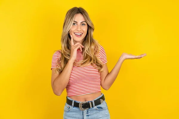 面白い若いです美しい女性身に着けているストライプTシャツ上の黄色の背景ホールドオープンパーム新製品素晴らしい提案 — ストック写真