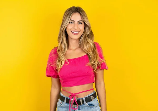 若い美しいブロンドの女性は 幸せとクールな笑顔で黄色のスタジオの背景にピンクのクロップトップを身に着けています 運のいい人 — ストック写真
