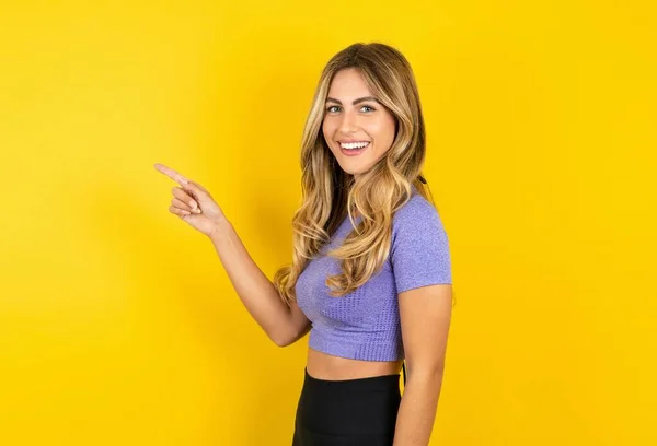 年轻漂亮的金发女人穿着运动服在黄色工作室背景上的侧面照片表明 手指空空如也 — 图库照片