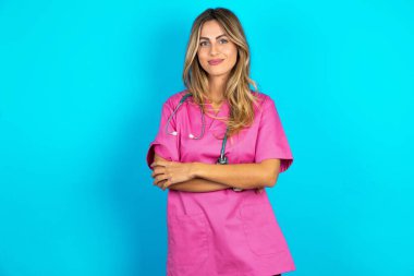 Düşünceli genç, düşünceli, güzel bir doktor. Mavi stüdyo arka planında duruyor. Kollarını kavuşturmuş, düşünceli bir ifadeyle ne yapacağını düşünüyor.