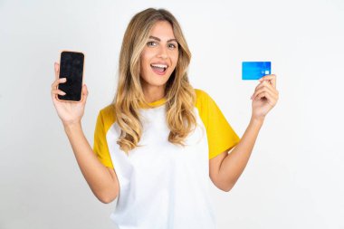 Beyaz arka planda futbol tişörtü giyen genç bir kadın banka hesabı açmış, elinde akıllı telefon ve kredi kartı varmış, gülümsüyor, online alışveriş başvurusu öneriyor.