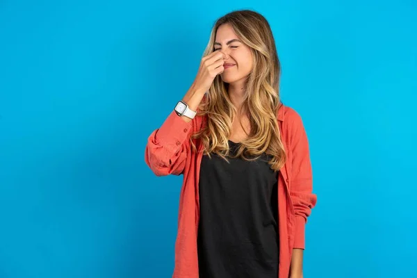 stock image young beautiful blonde woman standing over blue studio background smelling something stinky and disgusting, intolerable smell, holding breath with fingers on nose. Bad smell