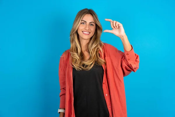 stock image young beautiful blonde woman standing over blue studio background smiling and confident gesturing with hand doing small size sign with fingers looking and the camera. Measure concept