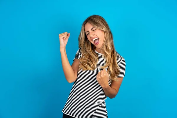 青のスタジオの背景に縞模様のTシャツを着た若い美しいブロンドの女性は非常に幸せと腕を上げ 笑顔と成功のために叫んで勝者のジェスチャーをやって興奮しています お祝いのコンセプト — ストック写真