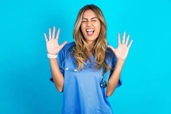 動機青いスタジオの背景の上に立つ若い美しい医師の女性は大声で笑い 面白い冗談や物語を聞き 満足して手のひらを上げる — ストック写真