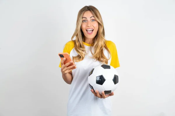 stock image Young beautiful woman wearing football T-shirt over white background holds mobile phone in hands and rejoices positive news, uses modern cellular