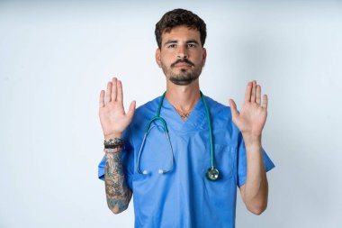 Beyaz arka planda cerrah üniforması giyen yakışıklı hemşire avuç içlerini kameraya çekiyor, jestleri durduruyor, duygularını kontrol etmesini ve gergin olmamasını istiyor.