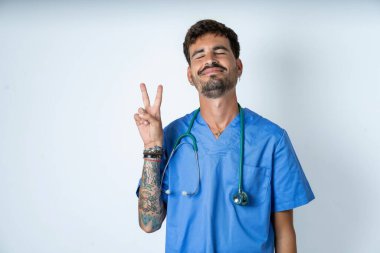 Beyaz arka planda cerrah üniforması giyen yakışıklı hemşire kameraya bakıp gülümsüyor ve zafer işareti yapıyor. İki numara..