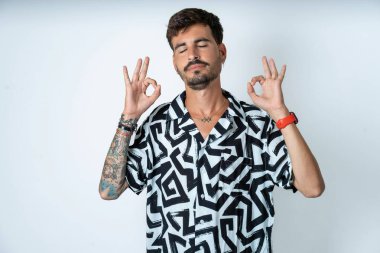 Yazlık gömlekli bir adam izole edilmiş beyaz arka planda dikiliyor rahatlıyor ve parmaklarıyla meditasyon yaparken gözleri kapalı gülümsüyor. Yoga konsepti.