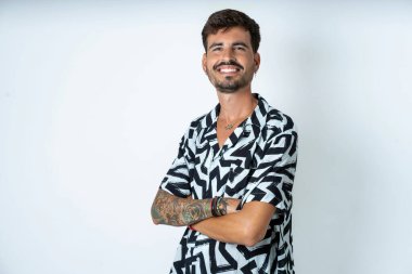 Yazlık gömlekli bir adam izole edilmiş beyaz arka plan üzerinde duruyor kollarını kavuşturup kameraya bakarken gülümsüyor. Pozitif kişi.