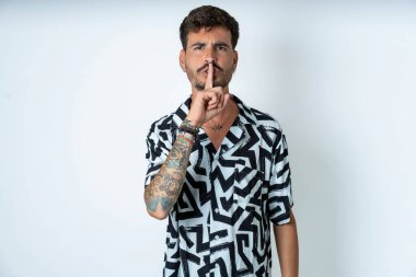 Yazlık gömlekli bir adam izole edilmiş beyaz arka planda duruyor sessiz bir jest yapıyor, parmaklarını dudaklarından üstün tutuyor. Sessizlik ve gizli konsept.