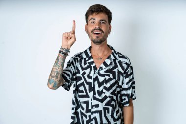 Baskılı tişört giyen genç beyaz bir adam parmağıyla işaret ediyor ve dahice düşüncelerden esinlenerek bakıyor, iyi bir fikir işareti gösteriyor, zekice bir çözüm düşünüyor.