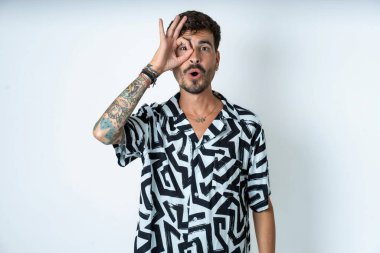 Baskılı tişört giyen genç beyaz adam şaşırmış yüz ifadesiyle şaşırmış, parmaklarının arasından bakan bir jest yapıyor. İnançsız ifade.