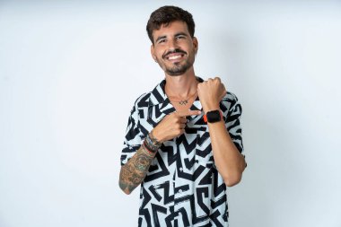 Yazlık gömlekli bir adam izole edilmiş beyaz arka planda dikiliyor. Aceleyle kol saatini işaret ediyor, sabırsızlıkla kameraya bakıyor ve rahat bir ifadeyle bakıyor.