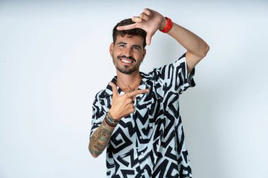 Yazlık gömlekli bir adam izole edilmiş beyaz arka planda duruyor ve elleriyle parmak çerçevesi yapıyor. Yaratıcılık ve fotoğrafçılık kavramı.