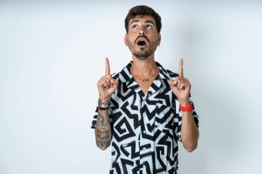 Baskılı tişört giyen genç beyaz adam şaşırmış ve şaşırmış bir şekilde yukarı baktı ve parmaklarıyla işaret etti ve kollarını kaldırdı..