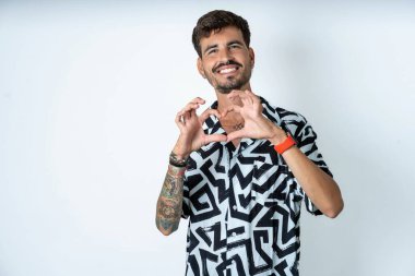 Yazlık gömlekli bir adam izole edilmiş beyaz arka planda durup aşkla gülümsüyor ve elleriyle kalp sembolü yapıyor. Romantik konsept.