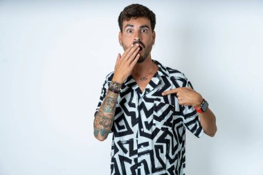 Yalıtılmış beyaz arka plan üzerinde duran dövmeli şoka uğramış bir adam şaşırtıcı bir şekilde aşağı bakıyor, ön parmağıyla bomboş, korkmuş bir model.