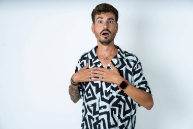 Yalıtılmış beyaz arka plan üzerinde duran dövmeli, korkmuş bir adam korkmuş bir ifadeyle ellerini göğsünde tutuyor, garip bir şey fark etmek için şaşırıyor, millet, sessiz tepki ve duygular..