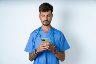 Heyecanlı, yakışıklı, cerrah üniforması giyen, beyaz arka planda göz kırpan, akıllı telefon kullanan, sosyal ağ haberlerini okuyan hemşire.