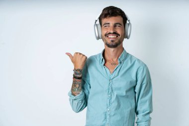 Beyaz arka planda turkuaz gömlek giyen yakışıklı genç adam kablosuz kulaklıkla ses kaydını dinliyor.